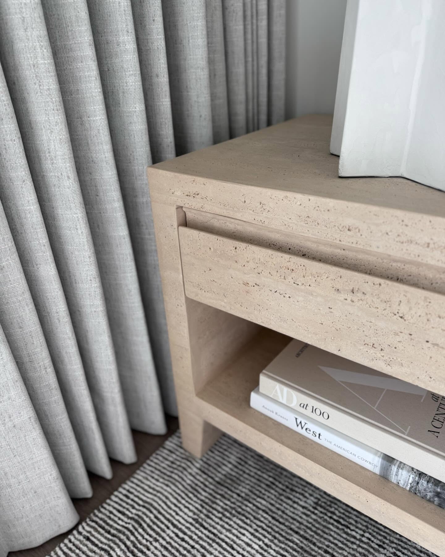 Travertine nightstand layered with design books beside softly draped linen bedding
