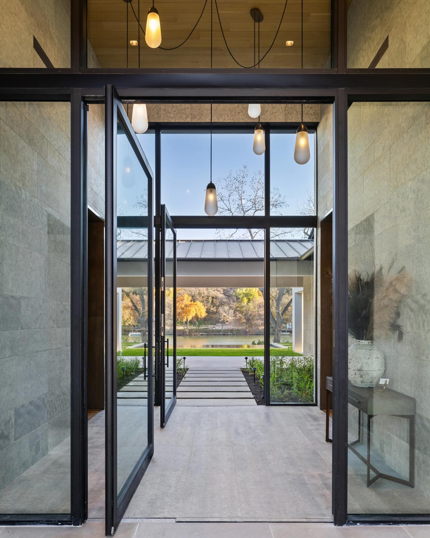 Double-height glass and steel entry with limestone walls opening to a lawn, pool, and lake framed by autumn trees