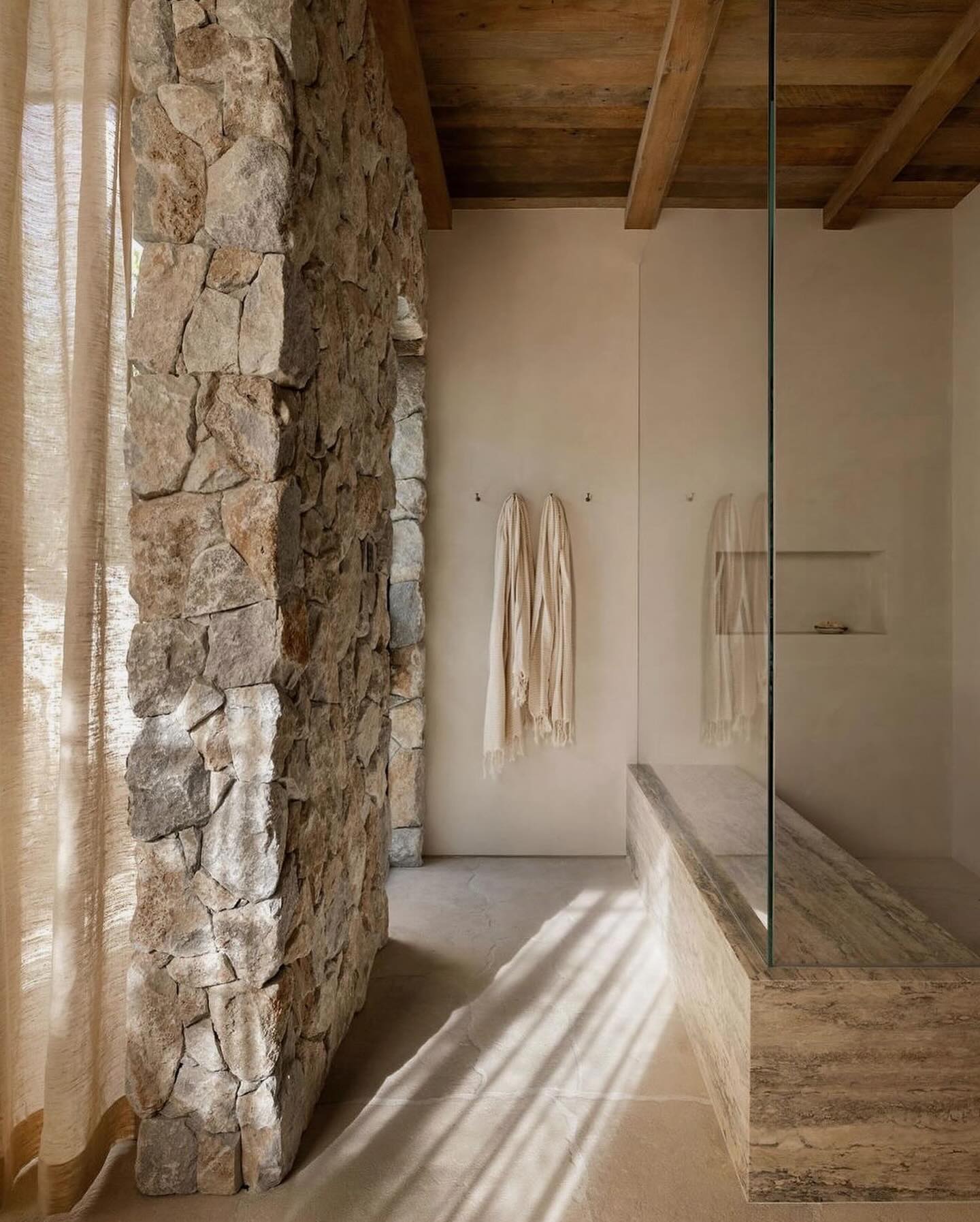 Spa bathroom with monumental fieldstone wall, timber beams, and warm raking sunlight