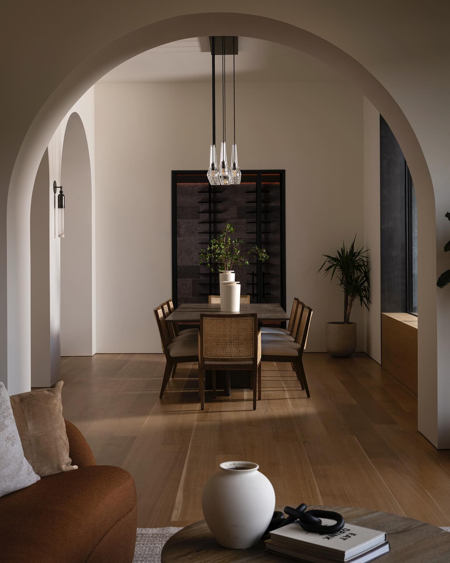 Dining room seen through a plaster arch, with glass globe pendants above the table and a full-height glass wine wall beyond